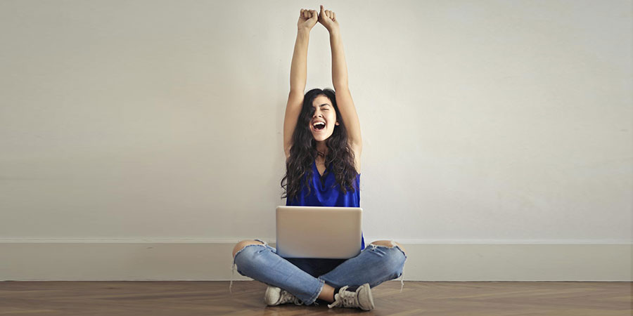 a woman wearing blue top and blue denim jeans sitting on the floor while raising her arms over her head with her laptop on her laps