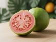 Halved guava with bright pink flesh and visible seeds next to whole guavas on a light wooden surface.