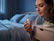 Person in a dim bedroom holding a melatonin bottle near a chamomile sprig on a bedside table.