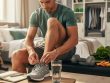 A man ties his athletic shoes next to a notebook and water in a bright home setting with workout and healthy food items nearby.